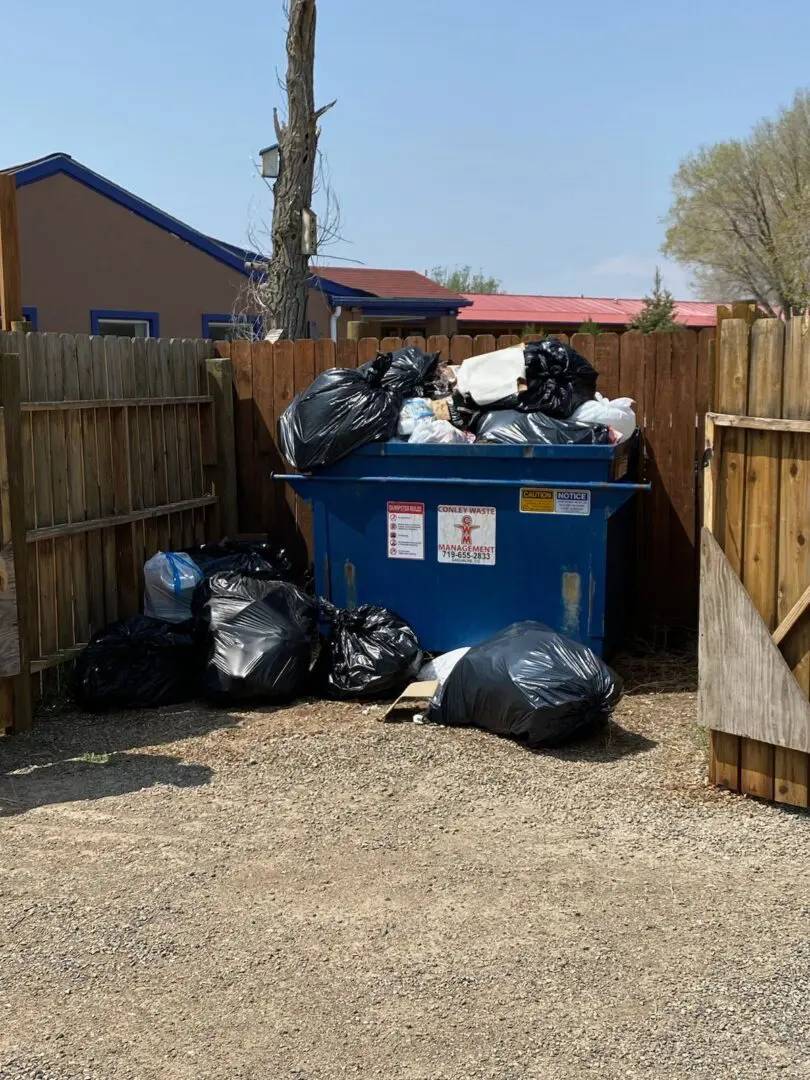 A dumpster full of trash bags in the middle of a fenced yard.